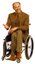 Gary sitting in his wheelchair in a jacket and tie gesturing with a smile.