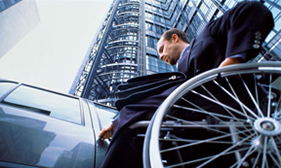 A businessman in a wheelchair wearing a suit next to a shiny car in front of a modern office building.