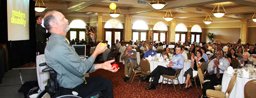 Gary Karp in a business suit, sitting in his wheelchair juggling three balls.